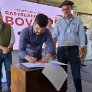 A imagem mostra um homem assinando um documento enquanto outros dois estão posicionados ao lado, olhando para ele. Ao fundo, há um painel indicando o tema do evento sobre rastreabilidade bovina. 