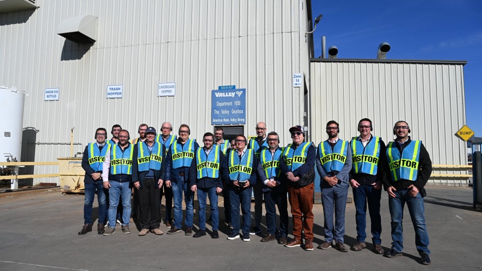 A imagem apresenta um grupo de visitantes posicionado em frente a um grande galpão industrial de paredes metálicas claras. Todos vestem coletes de segurança fluorescentes com a inscrição “VISITOR”.