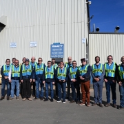 A imagem apresenta um grupo de visitantes posicionado em frente a um grande galpão industrial de paredes metálicas claras. Todos vestem coletes de segurança fluorescentes com a inscrição “VISITOR”.