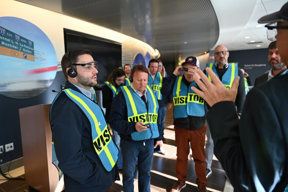 A foto mostra um grupo de pessoas em um ambiente interno. Todos usam coletes de segurança fluorescentes com a palavra “VISITOR”. Em primeiro plano, há uma pessoa gesticulando, conduzindo a apresentação.