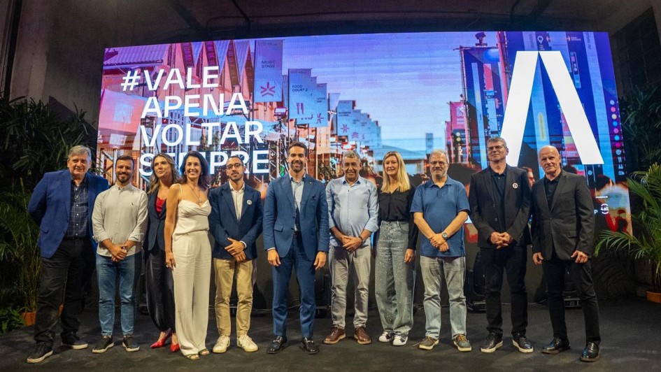 A foto mostra um grupo de pessoas alinhadas sobre um palco, posando para a foto, em frente a um grande painel digital colorido.