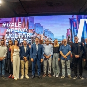 A foto mostra um grupo de pessoas alinhadas sobre um palco, posando para a foto, em frente a um grande painel digital colorido.
