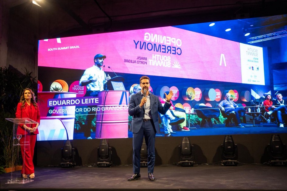 A foto mostra um palco com um grande telão ao fundo exibindo o nome “South Summit Brazil”, além de logotipos e imagens relacionadas ao evento. No centro do palco, o governador segura um microfone e faz uma apresentação.