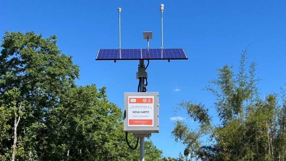 A foto mostra uma estação meteorológica automatizada instalada em área externa, cercada por vegetação verde e céu azul limpo.