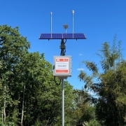 A foto mostra uma estação meteorológica automatizada instalada em área externa, cercada por vegetação verde e céu azul limpo.