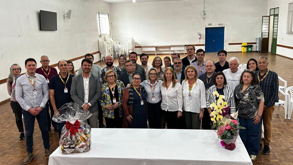 A imagem mostra um grupo de pessoas reunidas em pé, posando para a foto. Elas estão em uma sala grande, com uma mesa retangular com arranjos de flores na frente. 
