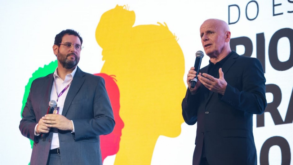 Dois homens em trajes sociais falam ao microfone no palco do evento. Ao fundo, painel colorido com silhuetas e texto sobre governo do Rio Grande do Sul. Cena formal e pública.