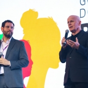 Dois homens em trajes sociais falam ao microfone no palco do evento. Ao fundo, painel colorido com silhuetas e texto sobre governo do Rio Grande do Sul. Cena formal e pública.