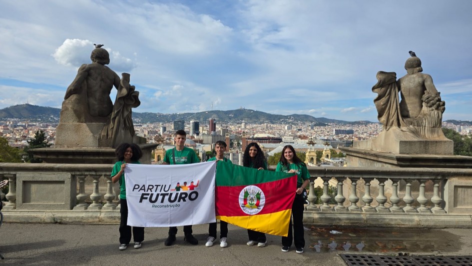 Cinco jovens estão em pé diante de um mirante com vista panorâmica de uma cidade com morros ao fundo. Eles seguram duas bandeiras: à esquerda, a do programa “Partiu Futuro Reconstrução” e, à direita, a bandeira do Rio Grande do Sul. Ao fundo, há esculturas de pedra voltadas para a paisagem urbana e o céu parcialmente nublado.