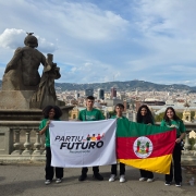 Cinco jovens estão em pé diante de um mirante com vista panorâmica de uma cidade com morros ao fundo. Eles seguram duas bandeiras: à esquerda, a do programa “Partiu Futuro Reconstrução” e, à direita, a bandeira do Rio Grande do Sul. Ao fundo, há esculturas de pedra voltadas para a paisagem urbana e o céu parcialmente nublado.