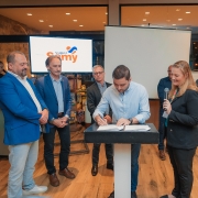 O vice-governador Gabriel Souza está assinando um documento sobre um púlpito, enquanto uma mulher ao seu lado segura um microfone. Outras seis pessoas, em pé, observam a ação.