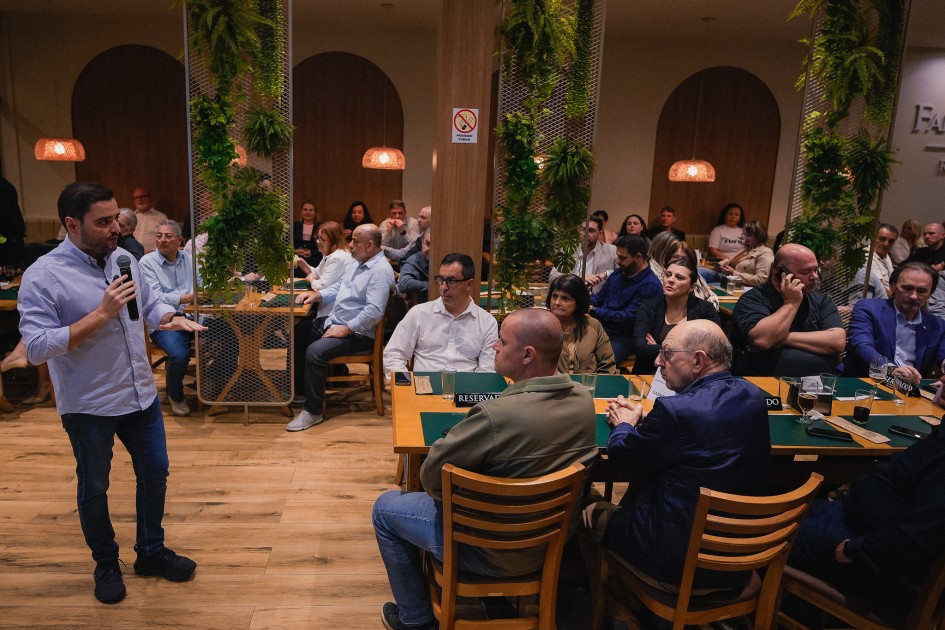 O vice-governador Gabriel Souza fala ao microfone diante de um grupo de pessoas sentadas em mesas de madeira em um ambiente interno. O público observa atentamente a apresentação. O local possui divisórias decoradas com plantas e iluminação amarela.