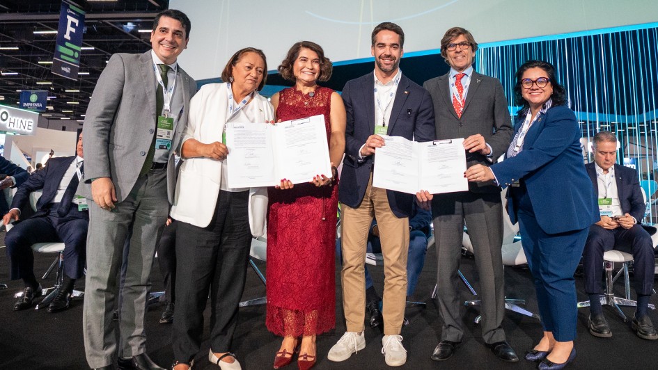 A imagem apresenta um grupo de seis pessoas em pé sobre um palco, segurando documentos abertos voltados para a câmera. Ao centro do grupo, está o governador Eduardo Leite.