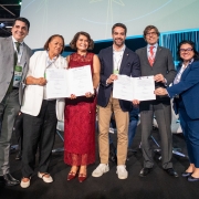 A imagem apresenta um grupo de seis pessoas em pé sobre um palco, segurando documentos abertos voltados para a câmera. Ao centro do grupo, está o governador Eduardo Leite. 