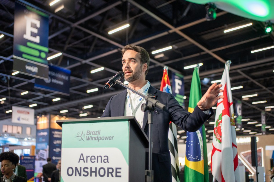 A foto mostra o governador Eduardo Leite em pé atrás de um púlpito com a inscrição “Brazil Windpower – Arena ONSHORE”. Ele fala ao microfone e gesticula com a mão.