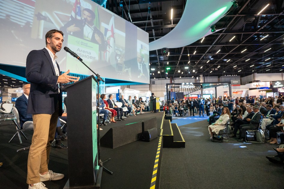 A foto mostra o governador discursando ao microfone em um palco, com um púlpito. À esquerda, há uma grande tela exibindo a imagem do orador em tempo real. No fundo do palco, várias pessoas estão sentadas em cadeiras, enquanto à frente há um público numeroso acompanhando a apresentação.