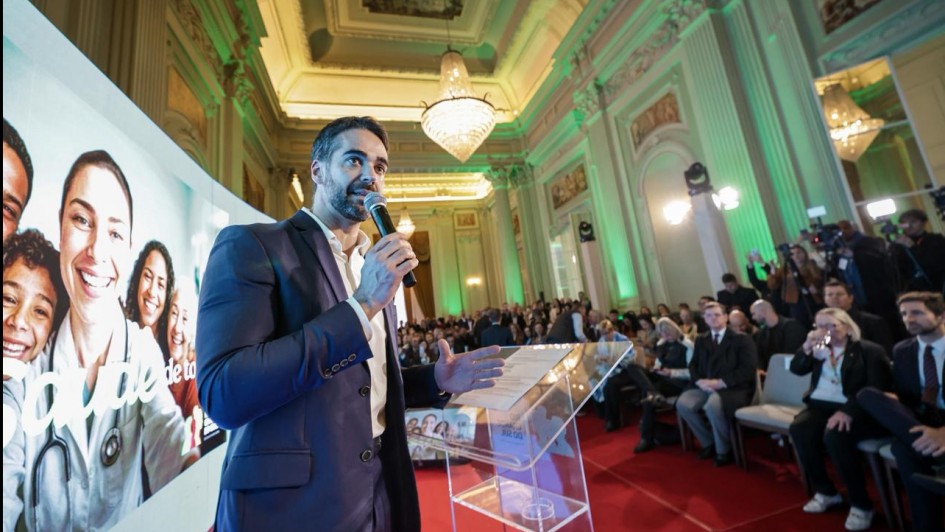 Evento formal em salão luxuoso, com homem de terno falando ao microfone num púlpito transparente. Ao fundo, exibe imagem de pessoas sorrindo, revelando tema de saúde ou comunidade. Público atento e presença da imprensa.