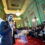 Evento formal em salão luxuoso, com homem de terno falando ao microfone num púlpito transparente. Ao fundo, exibe imagem de pessoas sorrindo, revelando tema de saúde ou comunidade. Público atento e presença da imprensa.