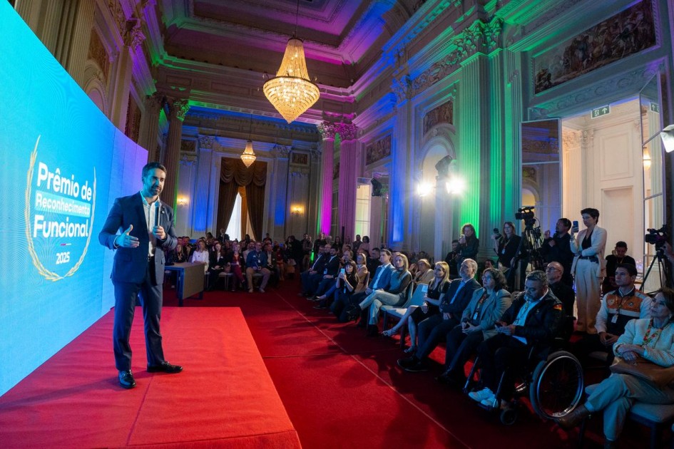 Foto em ambiente fechado, no Palácio Piratini. Leite aparece à esquerda do quadro, falando de pé ao microfone, à frente de um telão azul. À direita do quadro está a plateia sentada.