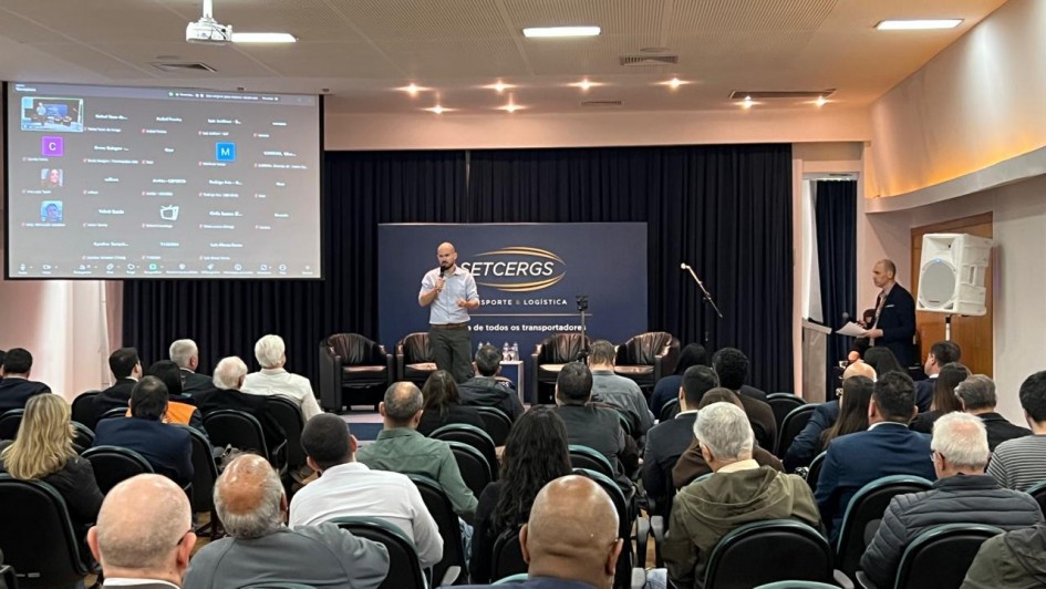 A imagem mostra um palestrante falando ao microfone em um auditório lotado. Do lado esquerdo da foto, há um telão.