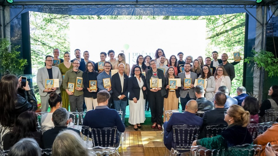 Uma foto de um Uma foto de um grupo grande de pessoas (cerca de 30, homens e mulheres) posando em um palco após receberem premiações. Eles estão de pé, sorrindo, e a maioria segura placas ou troféus retangulares com ilustrações. Eles são os vencedores do Prêmio Sema-Fepam de Jornalismo Ambiental 2025.

O palco é decorado com um grande painel de LED central que está totalmente iluminado com uma luz branca forte, e é emoldurado por painéis laterais que projetam imagens de folhagem verde, dando a impressão de natureza. Na frente do palco, há uma plateia sentada em cadeiras transparentes (de acrílico) e escuras. O ambiente é formal, mas com um toque de evento corporativo ou premiação.