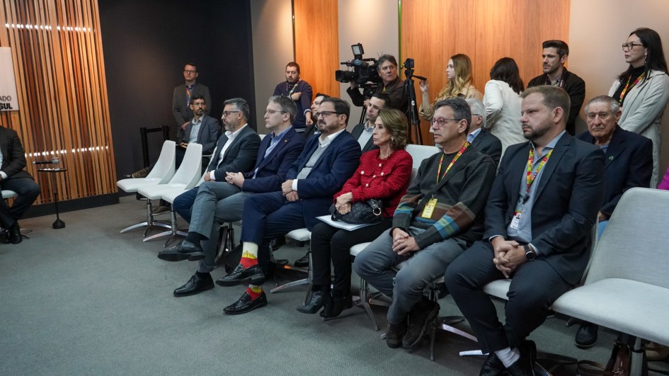 A imagem mostra uma plateia sentada em fileiras de cadeiras estofadas claras, focada em uma apresentação que não está visível. O ambiente tem um estilo moderno com paredes de painéis de madeira ripada e piso acarpetado cinza.
A maioria das pessoas está vestida com trajes de negócios. A fileira principal, no primeiro plano e meio-termo, é composta por aproximadamente nove pessoas, olhando para a frente.
Pessoas em destaque (Primeiro plano): Um homem à direita, usando um terno escuro e uma credencial colorida, está sentado com as mãos cruzadas. Ao lado dele, um homem de óculos veste uma jaqueta escura e uma camiseta com detalhes coloridos e também usa uma credencial. Entre eles e o homem de terno, há uma mulher de casaco de tweed vermelho e cabelos castanhos ondulados, segurando uma bolsa preta.
Outros na fileira: Seguem-se homens em ternos escuros ou blazers, alguns usando óculos. Um deles se destaca por usar meias amarelas e sapatos pretos.
Fundo: Na fileira de trás, há algumas pessoas em pé ou sentadas mais atrás. Entre elas, estão cinegrafistas com câmeras e microfones, e algumas pessoas em pé ao longo da parede, incluindo uma mulher de casaco claro à direita. Um homem de terno está sentado na extrema esquerda.
A cena retrata um público de profissionais em uma reunião ou evento.