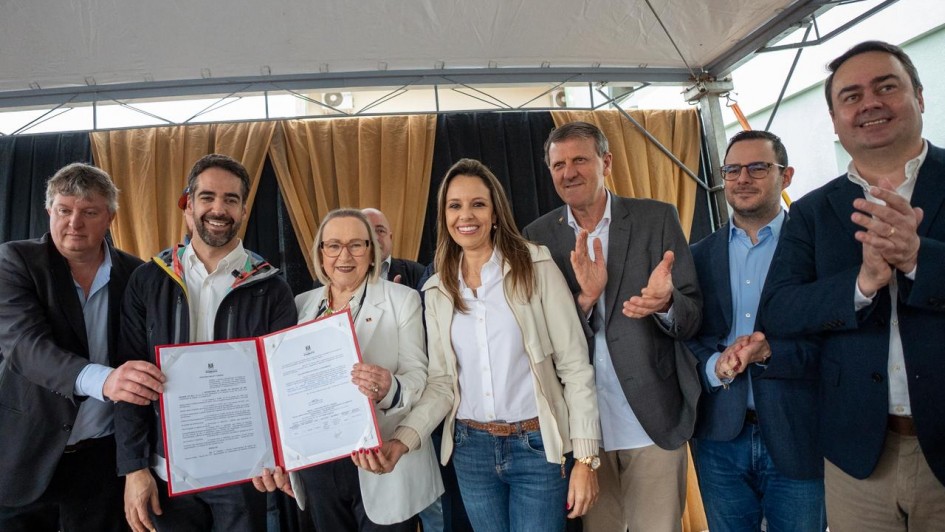 A foto mostra sete pessoas em pé, posando para a foto e sorrindo. O governador e a secretária Arita seguram e mostram um documento assinado.