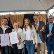 A foto mostra sete pessoas em pé, posando para a foto e sorrindo. O governador e a secretária Arita seguram e mostram um documento assinado. 
