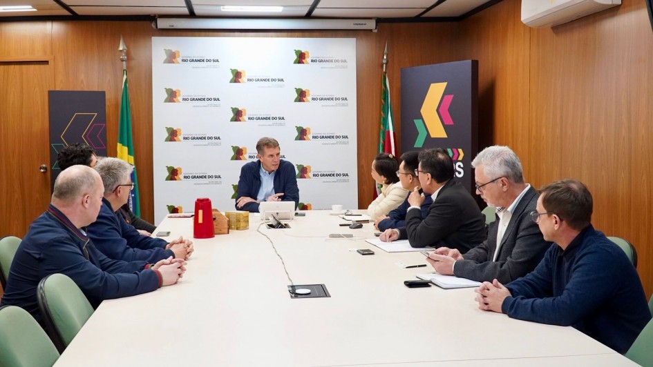 O secretário de Desenvolvimento Econômico, Ernani Polo, está reunido com um grupo de pessoas em torno de uma mesa retangular em sala de reuniões do governo estadual. O ambiente tem painéis de madeira, bandeiras do Brasil e do Rio Grande do Sul e o logotipo “invest.RS” ao fundo.