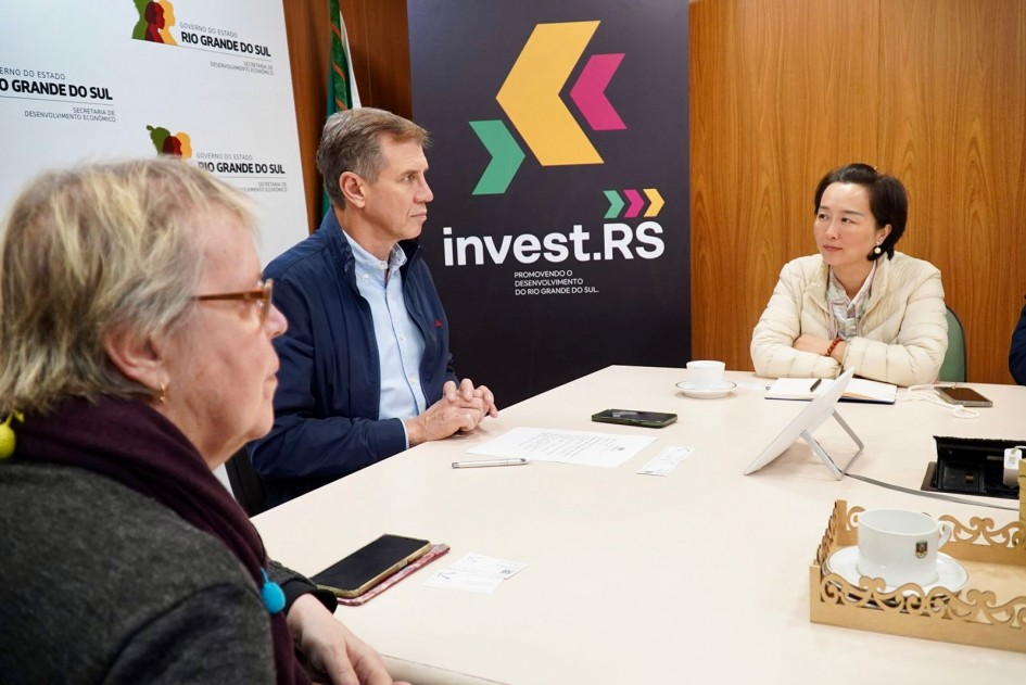 O secretário Ernani Polo aparece ao centro da imagem, sentado à mesa em reunião com uma mulher chinesa e outros participantes. Atrás dele há um painel com o logotipo “invest.RS – Promovendo o desenvolvimento do Rio Grande do Sul”. Sobre a mesa há xícaras, celulares e documentos.