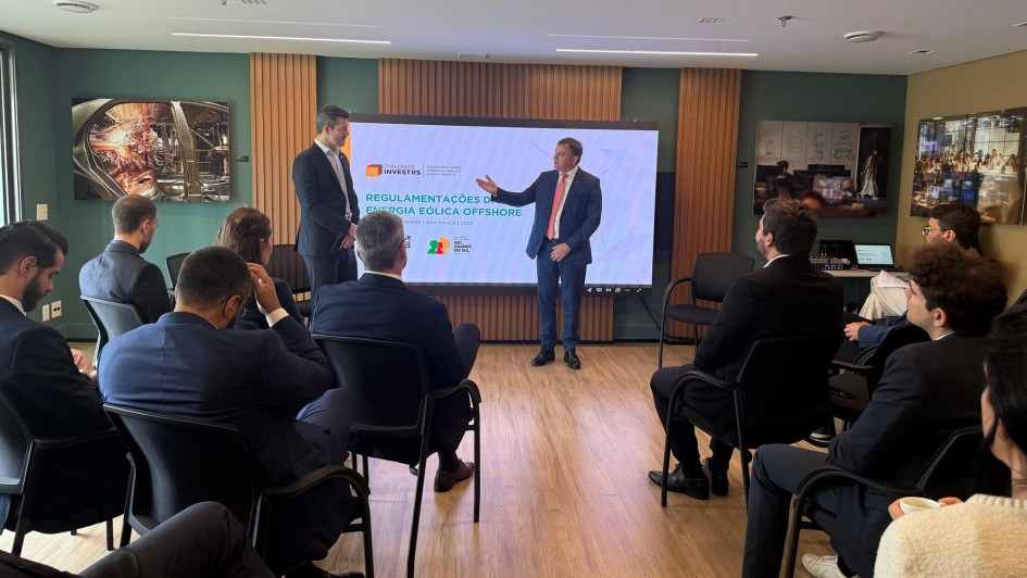 A imagem registra um evento ou reunião de negócios em um ambiente interno moderno, com piso de madeira, paredes em tons neutros (branco e marrom/verde musgo) e iluminação embutida.
No centro da sala, um grande telão exibe o tema da reunião: "REGULAMENTAÇÕES DE ENERGIA EÓLICA OFFSHORE" (Energia Eólica Marítima). Há logotipos associados, incluindo o da InvestRS e do Governo do Estado.
Oradores e Apresentação: Dois homens de terno escuro estão em frente ao telão. O homem à esquerda está de costas para a plateia, olhando para o orador. O orador principal, à direita do telão, está de frente para a plateia, gesticulando com a mão direita e sorrindo. Ele é branco, de cabelos claros e veste um terno azul-marinho com gravata vermelha.
Plateia: O público, composto majoritariamente por homens de terno escuro (representando empresários e especialistas), está sentado em cadeiras de escritório pretas, dispostas em fileiras laterais, de frente para os oradores e o telão. Eles estão focados na apresentação.
Detalhes do Ambiente: As paredes contêm painéis decorativos e algumas fotografias emolduradas com temas industriais/energéticos. Um dos painéis de parede é forrado em um tecido marrom escuro, e à direita, sobre uma mesa lateral, há equipamentos eletrônicos (como um notebook) e materiais de reunião.
A cena sugere uma discussão de alto nível sobre investimentos e regulamentação no setor de energia eólica offshore.