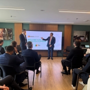 A imagem registra um evento ou reunião de negócios em um ambiente interno moderno, com piso de madeira, paredes em tons neutros (branco e marrom/verde musgo) e iluminação embutida.

No centro da sala, um grande telão exibe o tema da reunião: "REGULAMENTAÇÕES DE ENERGIA EÓLICA OFFSHORE" (Energia Eólica Marítima). Há logotipos associados, incluindo o da InvestRS e do Governo do Estado.

Oradores e Apresentação: Dois homens de terno escuro estão em frente ao telão. O homem à esquerda está de costas para a plateia, olhando para o orador. O orador principal, à direita do telão, está de frente para a plateia, gesticulando com a mão direita e sorrindo. Ele é branco, de cabelos claros e veste um terno azul-marinho com gravata vermelha.

Plateia: O público, composto majoritariamente por homens de terno escuro (representando empresários e especialistas), está sentado em cadeiras de escritório pretas, dispostas em fileiras laterais, de frente para os oradores e o telão. Eles estão focados na apresentação.

Detalhes do Ambiente: As paredes contêm painéis decorativos e algumas fotografias emolduradas com temas industriais/energéticos. Um dos painéis de parede é forrado em um tecido marrom escuro, e à direita, sobre uma mesa lateral, há equipamentos eletrônicos (como um notebook) e materiais de reunião.

A cena sugere uma discussão de alto nível sobre investimentos e regulamentação no setor de energia eólica offshore.