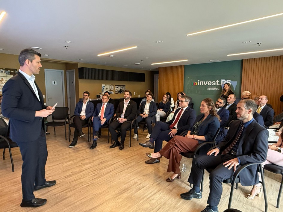 A imagem registra um evento ou reunião de negócios em um ambiente interno. O local possui piso de madeira, paredes em tons claros e uma parede de destaque no fundo revestida com painéis de madeira ripada.
Orador: No plano esquerdo, um homem de terno escuro está em pé, de perfil para a câmera, falando para a plateia. Ele é branco, tem cabelos e barba escura e segura um papel ou cartão nas mãos.
Plateia: O público está sentado em cadeiras de escritório pretas, dispostas em um semicírculo ou em fileiras laterais, preenchendo o restante do espaço. A plateia é composta por homens e mulheres vestidos em trajes de negócios.
Pessoas em destaque na plateia (Primeiro plano): Um homem à extrema direita está sentado, olhando para o orador. Uma mulher sentada logo atrás dele usa um blazer azul-marinho e calças marrons escuras.
Ambiente e Identificação: Na parede de fundo, à direita, há uma placa com o logotipo e o nome "invest.RS", identificando a sede do evento (provavelmente a InvestRS em São Paulo, conforme o contexto do nome do arquivo). Portas fechadas, que parecem levar a banheiros (indicado por símbolos de "homem" e "mulher"), estão visíveis na parede de fundo à esquerda.
A cena retrata um orador apresentando-se a um grupo de profissionais em um evento corporativo.
