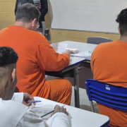 A imagem mostra quatro homens de costas, vestindo uniformes nas cores laranja e branco. Eles estão sentados em classes escolares, com papéis e canetas nas mãos. 