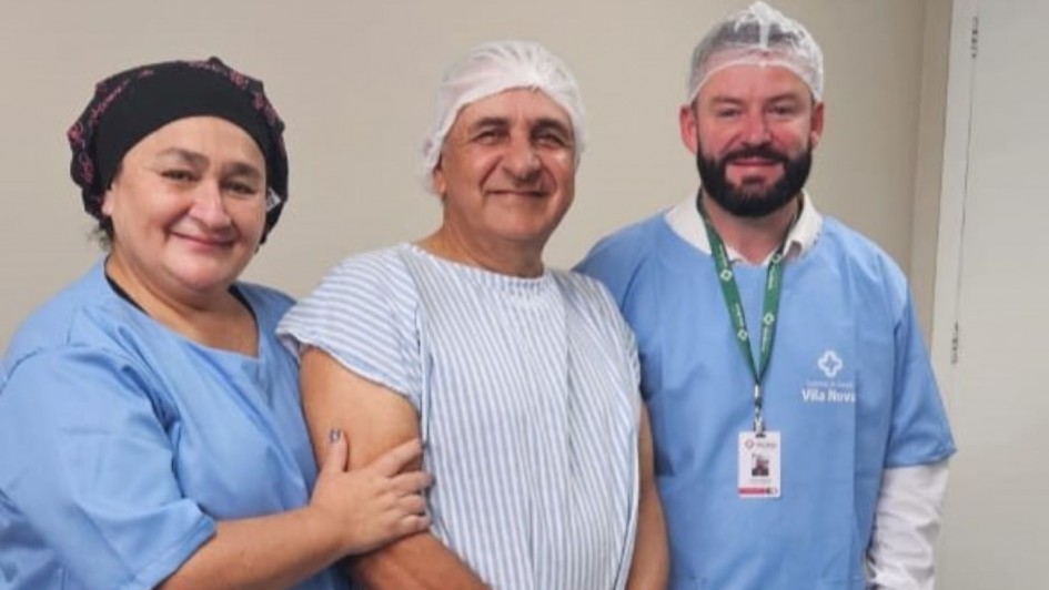 Dois profissionais de saúde e um paciente posam para a foto em ambiente hospitalar. Os profissionais vestem roupas e toucas azuis, enquanto o paciente, um homem, ao centro, de avental listrado e touca branca, sorri para a câmera. Todos estão em pé, diante de parede clara.