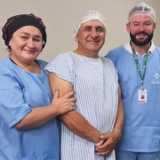 Dois profissionais de saúde e um paciente posam para a foto em ambiente hospitalar. Os profissionais vestem roupas e toucas azuis, enquanto o paciente, um homem, ao centro, de avental listrado e touca branca, sorri para a câmera. Todos estão em pé, diante de parede clara.