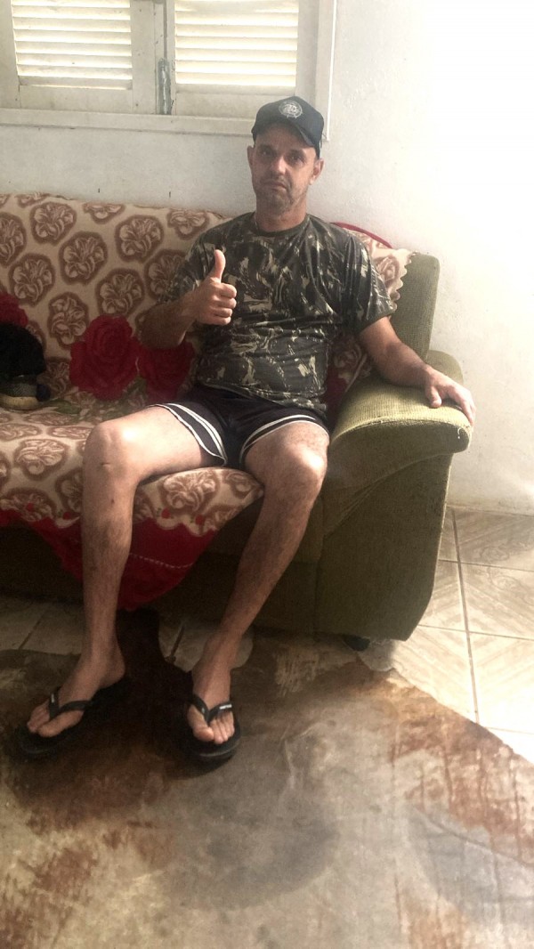 Homem sentado em sofá com estampa floral, usando camiseta camuflada, boné preto, shorts e chinelos, faz sinal de positivo com a mão direita. A foto é tirada em ambiente doméstico, com parede e janela ao fundo.