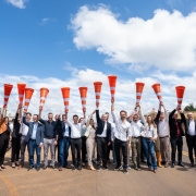 A imagem apresenta um grupo numeroso de pessoas alinhadas sobre o asfalto, segurando cones de sinalização laranja erguidos acima da cabeça. 