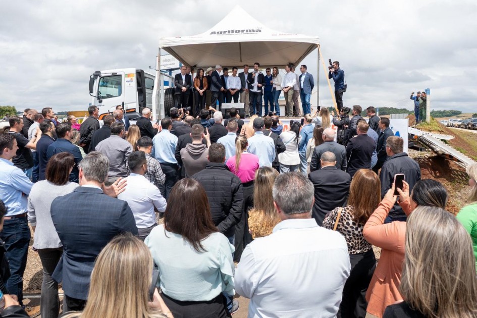 A foto mostra um evento realizado sob uma tenda branca montada sobre uma área pavimentada. No palco, o governador fala ao microfone e várias pessoas estão posicionadas lado a lado, enquanto um público numeroso acompanha em pé, voltado para a estrutura.