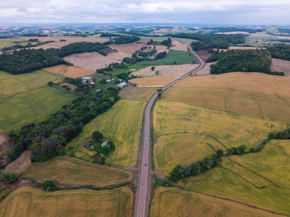 A foto aérea mostra uma rodovia asfaltada cortando uma paisagem rural com campos cultivados em tons de verde e dourado.