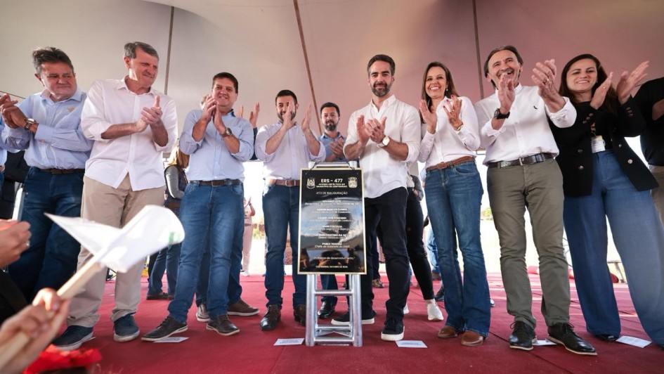 O governador Eduardo Leite aparece ao centro da imagem, em palco com outras autoridades. À frente deles, há uma placa de inauguração da ERS-477. O grupo aplaude o momento, em espaço coberto com piso vermelho.