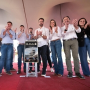 O governador Eduardo Leite aparece ao centro da imagem, em palco com outras autoridades. À frente deles, há uma placa de inauguração da ERS-477. O grupo aplaude o momento, em espaço coberto com piso vermelho.
