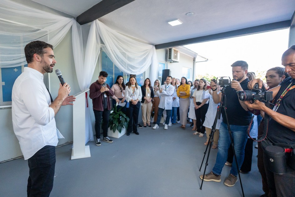 O governador Eduardo Leite, à esquerda, fala ao microfone diante de um grupo de profissionais de saúde e imprensa. Ele está em área coberta, decorada com tecido branco, e os ouvintes estão posicionados de frente para ele, alguns registrando o momento com câmeras.