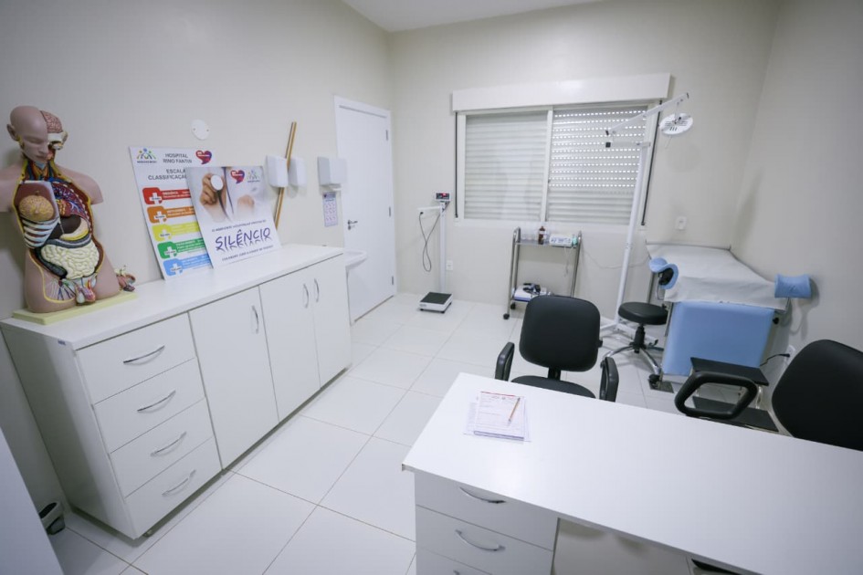 Imagem do interior de um consultório do Hospital Rino Fantin, com móveis brancos, mesa de atendimento, cadeiras, macas e equipamentos médicos. Sobre o balcão, há um modelo anatômico do corpo humano e cartazes informativos sobre classificação de risco e silêncio hospitalar.