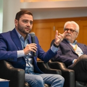 Dois homens sentados em poltronas pretas participam de conversa formal. Um fala ao microfone com expressão séria, o outro observa atento. Ambos usam blazer azul e jeans. Ambiente interno e profissional.