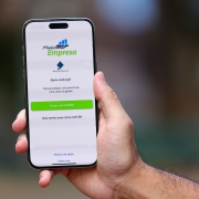 A imagem mostra uma mão segurando um smartphone. Na tela do celular aparece um aplicativo chamado Minha Empresa.