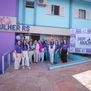 A foto mostra a fachada de um prédio pintado em tons de azul e lilás, com uma placa grande que diz “SER MULHER RS – Serviço Especializado de Referência à Saúde da Mulher”. Em frente à entrada, há um grupo de pessoas organizadas para uma foto coletiva, vestindo camisetas roxas.