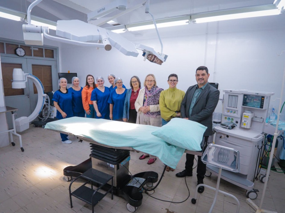 A imagem apresenta uma sala cirúrgica equipada com mesa de cirurgia, lâmpadas cirúrgicas suspensas, monitor e outros equipamentos médicos. Ao fundo, um grupo de pessoas está posicionado para foto, algumas vestindo roupas hospitalares azuis e toucas.