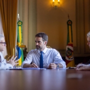 Foto de uma reunião num ambiente fechado. Dois homens e uma mulher aparecem sentados à mesa. Leite está na cabeceira, de frente para a câmera, olhando para o homem à esquerda, que fala.