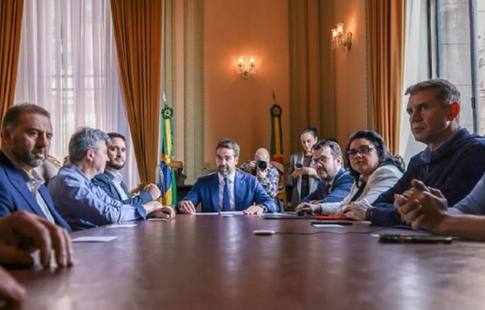 O governador Eduardo Leite aparece ao centro da mesa, em reunião no Palácio Piratini, com secretários e representantes de instituições participantes do edital de hidrogênio verde. A sala tem cortinas douradas, janelas amplas e bandeiras do Brasil e do Rio Grande do Sul ao fundo.